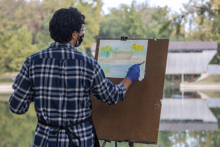 A student paints a scene of a covered bridge and reflecting pond at Mill Race Park.